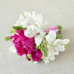 A petite, elegant floral piece rests on a pale beige, linen-textured cloth, the soft weave of the fabric providing a gentle, almost rustic frame for the colours. Dominating the centre is a sumptuous fuchsia to magenta flower, reminiscent of a small peony or garden rose, its many ruffled petals tightly cupped yet generously layered, giving a sense of plush depth and tenderness. Clustered closely around this focal bloom are bright white alstroemeria, their narrow, slightly curved petals tipped and streaked with maroon and deep burgundy markings that resemble fine calligraphy on porcelain. Sprigs of light, vibrant green buds and slender foliage peek between the petals, bringing in a fresh, meadow-like feel and stretching the eye along the length of the arrangement. The stems are bound together with a glossy, vivid pink satin ribbon, wound smoothly and neatly, its sheen catching the light and mirroring the saturated tone of the central bloom. The lighting is calm and diffused, with subtle shadows adding dimension to the petals and emphasising the texture of the background. This compact design has the refined scale of a boutonnière or wrist corsage, ideal for a bridegroom, bridesmaid, or prom guest heading out from Golders Green station towards a celebration in Temple Fortune or Finchley. The overall mood is one of modern romance and effortless charm, like something a skilled local florist has hand-tied just moments before delivery, ensuring the flowers arrive at their freshest for a meaningful occasion shared with close friends and family.