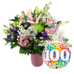 Set against a clean, pure white background, this jubilant floral display feels like a moment of celebration frozen in time. A matte pink, ribbed ceramic vase anchors the design, its soft blush tone echoing the pastel houses and café fronts you might pass along Golders Green Road. From this vase, a generous bouquet rises in a loose, rounded shape, overflowing with fresh flowers that look as though they have just been conditioned by an experienced florist. At the heart of the arrangement, large pink lilies unfurl their star-shaped petals, while several green-tipped buds reach upward, hinting at more blossoms to come. Nestled around them, layers of light pink and dusty rose-coloured roses create a plush, romantic core. Deep purple lisianthus stems thread through the roses, their silky petals adding depth, while slender sprigs of lavender-toned asters with tiny yellow centres lend a soft, meadow-like charm. Bright lime-green button chrysanthemums pop forward, their tight, spherical blooms giving a playful texture that contrasts with the airy spray of white baby's breath drifting through the design. Rich green foliage frames the composition, its leaves providing a cool, fresh backdrop to the warmer tones. To the lower right of the vase, partially overlapping the bouquet, a shiny round foil balloon glistens with a full surface of multi-hued confetti dots - jubilant specks of red, orange, yellow, blue, and green. Dominating the balloon is the large white number '100', slightly raised in effect, announcing a landmark birthday or centenary. The overall scene evokes the warmth of a family gathering in Golders Green, celebrating a loved one's 100 years with flowers that feel both modern and timeless.