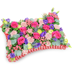 As a florist working locally around Golders Green and often delivering to services near Golders Hill Park and the tube, I picture this rectangular pillow tribute presented from a slightly raised, oblique viewpoint on a pristine white ground. A plush carpet of hot pink roses dominates the surface, each bud or gently opening bloom chosen for symmetry and colour intensity. Interspersed are pale pink spray carnations, their frilly, shell-like petals softening the composition and creating a comforting, tactile rhythm. Scattered pockets of purple lisianthus add cool contrast and a sculptural bell shape, while cheerful lemon-yellow ranunculus scatter luminous points that feel like small suns across the cushion. Sprays of silver-blue Eryngium bring an unexpected, spiky counterpoint that lifts the arrangement and adds interesting silhouette against the greenery. Rounded eucalyptus leaves with their muted, silver-green sheen nestle among darker foliage, lending a herbal scent that balances the rose sweetness. The whole piece sits upon a pleated, deep-red satin ribbon base that frames the floral field and gives the pillow its defined, elegant edge. Light falls softly across the petals, revealing velvety textures and subtle tonal shifts that comfort the eye. Intended as a funeral pillow or memorial cushion, the arrangement speaks of remembrance and celebration of a life-warm, tender and uplifting rather than solely mournful. At Flowers Golders Green we craft these tributes with respectful attention to scent, proportion and placement, available for careful local delivery across Golders Green and nearby Finchley Road.