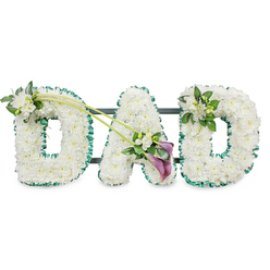 A compassionate, handcrafted memorial piece spelling out DAD in bold, three-dimensional letters sculpted from tightly clustered white chrysanthemums and similar small white flowers, producing a smooth, cushiony texture that reads as pure and reverent. Each letter is edged with a finely folded deep green satin ribbon that frames the white blooms with a fresh, natural contrast and a slight satin gleam under gentle light. The letters are mounted on a dark, unobtrusive frame-horizontal supports peek from behind-so the tribute stands upright for viewing at a funeral, memorial or graveside. Thoughtful floral touches bring movement and focus: a petite spray of white freesia and glossy foliage nestles into the upper left curve of the first D; two long green stems sweep diagonally across toward the A, where a tasteful asymmetrical cluster combines more white freesia, verdant leaves, and two striking deep-purple calla lilies whose trumpet forms introduce a deeper, dignified tone. The right-most D receives a smaller, mirrored grouping of freesia and greenery, balancing the piece without overpowering the message. The overall feeling is quiet and consoling, the faint perfume of freesia mingling with the soft, papery scent of chrysanthemum. As a local florist in Golders Green and surrounding streets like Finchley Road and North End Road, I often suggest this classical palette of white, green and a single dark accent for families honouring a father with a simple, heartfelt statement.