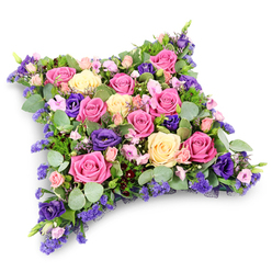 A luxurious rectangular floral cushion, finished with softly rounded corners, fills the frame like a bed of petals, its surface completely covered in roses and seasonal blooms. The colour palette moves gently from rich fuchsia and rose-pink through soft blush and cream to deeper tones of purple, creating a comforting harmony that feels both elegant and heartfelt. Large roses, some fully open and others just beginning to unfurl, form the heart of the tribute: vivid pinks and delicate creamy yellows mingle, their layered petals catching the light in subtle highlights and shadows. Between the roses, deep royal-purple lisianthus blooms add a velvety, almost satin texture, drawing the eye inward and providing a sense of depth, like twilight gathering over a quiet corner of Golders Green. Fine clusters of tiny lavender and lilac flowers - likely statice or limonium - nestle into any remaining spaces, so there is no gap left unfilled, giving the arrangement a plush, cushioned appearance. Around the flowers, rounded eucalyptus leaves in a soft silvery green curl and overlap, while darker, more feathery foliage peeks through to structure the design and create contrast. The tribute is photographed against a pure white background with a gentle shadow beneath, making the cushion appear as if it is resting lightly on a table in a calm chapel or funeral home near Golders Green Crematorium. Every detail communicates care, compassion, and professional craftsmanship, making this floral pillow a fitting choice for families in Golders Green who wish to honour a loved one with a peaceful, beautifully composed final gesture.