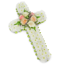 When silence needs a voice, this white chrysanthemum cross speaks with calm assurance. The structure is generously built from small, button-style white mums so closely packed they read like a soft, snowy blanket stretched into the familiar cross silhouette; the subtle drop shadow against the bright studio background gives the piece a gentle, sculptural presence. Nestled at the centre where vertical meets horizontal, a compact bouquet of fully opened roses introduces gentle warmth - petals in pale peach, creamy ivory and a tender blush of pink - each bloom selected to convey enduring love and tenderness. Wisps of fern add elegant, green filigree and a natural movement that contrasts the cross's solid form, while tiny lavender-hued filler flowers provide a cooling counterpoint to the roses' warmth. A slim lime-green ribbon trims the perimeter, the color brightening the white without breaking its serenity; it's tucked neatly so that it peeks out like a thoughtful accent rather than a flourish. You can almost sense the texture under your fingertips: the fine, cushiony mums, the silky rose petals, the soft serrated fronds. This arrangement is made with reverence for memorial traditions common in our neighbourhood - the kind of tribute families in Golders Green, near Finchley Road and Temple Fortune, choose for funerals, memorial services or to be laid at a graveside - a composed and comforting emblem of faith, remembrance and respect.