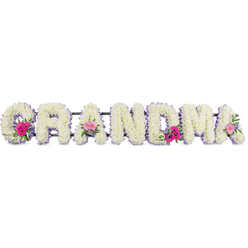 This image shows a beautifully crafted funeral flower tribute that spells out the word GRANDMA in large, upright letters, each one luxuriously covered in pristine white chrysanthemums. The blossoms are densely arranged so that no gaps in the letters are visible, forming a soft, cushion-like surface with countless little petals overlapping like feathers. Around the edge of every letter runs a slim lilac ribbon, the pastel purple trim giving each shape a clear outline and adding a calm, graceful contrast to the snowy white blooms. Three focal clusters of colour are woven into the design: on the first G, one of the central As, and the last A, compact posies of bright pink gerbera daisies and soft pink roses sit proudly on top of the chrysanthemums. These accent flowers create joyful bursts of pink against the white canvas, while sprigs of fresh foliage and small leaves lend touches of green that feel soothing and natural, like a walk through Golders Hill Park on a quiet afternoon. The tribute lies on a flat surface, likely prepared by experienced florists at Flowers Golders Green before a service or family farewell. The overall mood of the piece is tender and reverent-an elegant floral message that feels both strong and gentle, perfectly suited to honouring a grandmother whose presence once warmed family gatherings in Golders Green, Temple Fortune, or nearby streets. The composition balances purity, softness, and subtle colour, capturing the essence of remembrance and enduring love.