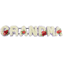 Set against a clean white background, this carefully composed funeral flower arrangement spells GRANDMA in large, bold letters, like a quiet statement of love laid in a Golders Green chapel. Each letter is built entirely from pristine white cushion chrysanthemums, packed closely together so their tiny, rounded petals create a soft, plush texture that looks almost like fresh snow. A delicate band of pale lavender or light purple ribbon traces the outline of each letter, lending a gentle glow of colour and a sense of serenity reminiscent of an early morning walk through nearby Golders Hill Park. Rich accents of red flowers are thoughtfully placed to punctuate the tribute. On the letter G, a lush spray of deep red roses curves into the shape, joined by a unique red-and-white variegated calla lily that adds a touch of individuality, as if capturing one of grandma's favourite blooms. The first A carries a single, heart-shaped red anthurium at its base, its glossy surface and sculptural form standing out against the cushiony white petals. At the centre, the M is enhanced with another cluster of velvety red roses and a second red anthurium, while the final A is finished with a solitary anthurium, completing the visual rhythm from start to finish. Subtle greenery and a dark green supporting frame hold the letters in place, giving the piece a steady, reassuring presence. This personalised floral lettering is a touching sympathy tribute, ideal for funeral services or a cremation near Golders Green, offering a graceful way to say goodbye to a much-loved grandmother with beauty, dignity, and quiet affection.