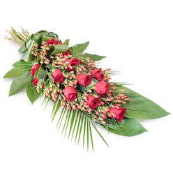 A refined funeral spray of red roses and berries rests on a crisp white background, its diagonal placement giving the arrangement a sense of gentle motion and quiet ceremony. At the top left, the stems are gathered tightly and wrapped in a smooth green leaf, creating a structured handle that feels both practical and beautifully finished. From that point, a river of deep crimson roses pours along the length of the design, each bloom with velvety petals and subtle variations in size and openness, from shy buds to generous, full flowers. Nestled among them are clusters of pinkish-red Hypericum berries, their small, rounded forms catching the light and softening the intensity of the roses with a slightly lighter red hue. Around this floral core, a rich collar of foliage spreads out: broad, glossy dark green leaves provide a steady, comforting backdrop, while airy palm fronds arc gracefully from the centre and lower right edge, stretching the spray into a long, elegant outline. Slender, twig-like stems and finer greenery thread through the base, adding the kind of layered texture a seasoned florist in Golders Green would build by hand at the workbench just off Golders Green Road. The colour palette of deep red and fresh green, framed by the stark white background, creates a striking yet respectful composition. This piece feels perfectly suited to rest beside a coffin in a local chapel or to be delivered to a family home near Hampstead Garden Suburb, offering a quiet expression of sympathy, remembrance, and enduring affection.