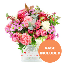 A richly detailed romantic bouquet fills the frame, arranged with the care of an experienced local florist familiar with Golders Green's cosy homes and leafy streets. The flowers rise from a clear, square glass vase, set against a pristine white background that accentuates every tone of pink, purple, and green. Several large roses in shades of fuchsia and soft blush pink form the focal point, their petals layered and slightly curled at the tips, some brushed with deeper magenta that adds drama and depth. Nestled beside them are ample carnations in warm coral and peach-pink, their ruffled petals creating a soft, almost feathery texture that feels both generous and comforting, like a hug sent in floral form. Cheerful lavender and purple daisy-like chrysanthemums with electric green centres pop up throughout the design, bringing a sense of movement and playfulness, as though they were picked on a bright morning stroll towards Golders Green station. The bouquet is wrapped visually in lush greenery: rounded eucalyptus leaves in silvery green, glossy darker foliage, and subtle touches of reddish-brown leaves that lend a hint of seasonal romance. The vase itself, with its clean lines and transparent sides, is decorated around the base by a white band sprinkled with vivid red hearts and the handwritten-style phrase all my love, quietly reinforcing the sentimental message. In the lower right corner of the image, an orange circular badge announces VASE INCLUDED in bold white letters, signalling that this is a complete, ready-to-display gift. The arrangement feels ideal for birthdays, anniversaries, or a surprise same-day delivery around Temple Fortune or Hampstead Garden Suburb, radiating warmth, affection, and modern elegance.