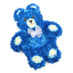 A meticulously shaped floral teddy bear rests against a bright white background, its entire form crafted from tightly packed blossoms that suggest the softness of a well-loved toy. The bear's body is cloaked in vivid royal blue flowers, each miniature head carefully aligned so that the petals create a seamless, velvety surface from rounded ears to outstretched paws. This rich blue colour is deep and soothing, evoking calm skies over the tree-lined streets of Golders Green on a quiet afternoon. Creamy white and pale lemon blooms highlight the bear's inner ears, muzzle, and oval paw pads, their lighter tones gently lifting the design and giving definition to the familiar teddy bear shape. The face is gently expressive: two rounded, dark amber eyes appear to shine with a soft reflection, while the small black triangular nose and curved stitched-like mouth offer a tender, reassuring smile. Around its neck, a sheer white organza bow with ruffled edges sits neatly in place, its translucent loops catching the light and adding a whisper of ceremony. Every curve of the bear's body feels intentionally sculpted, as though the flowers have been brushed into place rather than wired and pinned. This tribute carries a quiet emotional weight, perfectly suited to a child's funeral, a baby boy's remembrance, or a thoughtful sympathy gift to be delivered to a home near Golders Green Road or a chapel by the crematorium. The design communicates innocence, love, and gentle farewell, turning living petals into a comforting symbol of presence and memory.