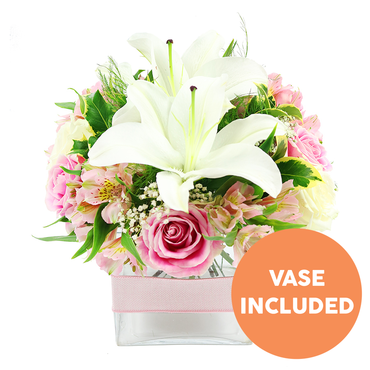 A stylish, ready-to-display bouquet is shown against a crisp white backdrop, spotlighting a graceful mix of white lilies and pink blooms designed in a compact glass vase. Two prominent white Oriental lilies rise at the heart of the arrangement, their broad petals elegantly curved and their dark reddish-brown stamens standing tall, drawing the eye to the centre. Embracing these lilies is a generous collar of blush to medium-pink roses, some just beginning to open with tightly coiled centres, others fully blossomed with soft, layered petals that suggest tenderness and romance. Between the roses, slender stems of pink and white alstroemeria add a playful flicker of colour, their petals lightly freckled with yellow, while tiny stars of white gypsophila float like mist around the larger flowers, making the bouquet appear airy despite its structured dome shape. Various shades of green foliage provide a cool, refreshing backdrop: glossy leaves catch subtle reflections, and finer, fern-like greens create delicate shadows, giving depth as if the arrangement were sitting by a bright window in a Golders Green flat near Temple Fortune. The flowers are carefully gathered into a low, square clear glass vase, where the stems are neatly cut and immersed in clear water, conveying freshness and expert craftsmanship from a local North West London florist. A pale pink satin ribbon is wrapped smoothly around the middle of the vase, adding a refined decorative band that hints at celebration and care-ideal for a dining table, bedside, or a same-day delivery to a colleague's office on Golders Green Road. In the lower right corner of the photo, a bold orange circular emblem with white capital text states VASE INCLUDED, making it clear the chic container accompanies the floral design.