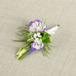 This boutonniere is a small, deliberate composition that celebrates subtle colour and careful touch, photographed on neutral linen that softens the scene. Center stage are two lisianthus blooms-snow-white petals tipped with a cool violet hue-whose layered, ruffled faces suggest a velvet softness beneath the fingertips. Interspersed among them, tiny sprays of baby's breath float like froth, lending a delicate brightness that lifts the arrangement. A few slender, pale buds arc outward, adding a sense of forward movement and promise. Beneath the blossoms, compact clusters of grape hyacinth introduce a deeper blue-violet counterpoint: small, bead-like florets that anchor the lower plane and provide visual contrast without overpowering the lisianthus. Wisps of fern add an airy, green filigree that frames and softens the silhouette, their texture giving an almost woodland quality to the boutonniere. The stems are bound with a precise white satin ribbon, its smooth sheen mirroring the linen beneath and securing the piece for pinning. In soft light-the kind you might find in a south-facing front room near Golders Hill Park-the tactile interplay of silk-fine petals, feathery filler, and crisp ribbon becomes very apparent, as does the faint, sweet scent that accompanies such fresh blooms. Crafted with local care by Flowers Golders Green for weddings, portraits, or memorable occasions in the area, this boutonniere feels like a small, thoughtful poem of spring designed to be noticed in quiet moments.