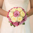 A soft, domed bridal bouquet fills the frame, a tender gathering of creamy white roses and blush-to-magenta pink blooms that seem to glow with their own inner light, as if photographed near a bright window above Golders Green station. Each rose is tightly petalled, unfurling in plush spirals that create a sense of richness without excess, the ivory blossoms blending seamlessly into the pinks so that the colours drift into one another like a quiet sunrise. Dotted delicately between the larger flowers are airy tufts of white baby's breath, their tiny clustered florets adding a cloud-soft texture that lightens the overall composition. At the base, soft green leaves gently cradle the bouquet, visible only in discreet glimpses, giving the hand-tied design a fresh, just-cut-from-the-garden feeling. The bouquet forms a near-perfect round, balanced from every angle, suggesting the practiced eye of a florist who works often with brides marrying in nearby Hampstead or along Finchley Road. There is a sense of stillness and serenity in the arrangement, as though the roses are pausing in a breath before the vows are spoken. You can almost sense the cool, satin touch of the petals and inhale the romantic fragrance-a gentle mix of classic rose, spring air, and the faint sweetness that lingers after rain on the pavements of Golders Green. Designed for a bride who loves elegance without ostentation, this bouquet captures that private, fluttering excitement just before walking down the aisle.