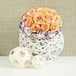 This image presents a lovingly curated bouquet that balances modern simplicity with romantic charm: a dense, dome-shaped cluster of apricot and soft-peach roses, roughly two to three dozen strong, each bloom showing a pleasing mix of fully opened faces and tighter buds. Encircling the roses at their base are white Lisianthus flowers, their crepe-like petals tipped in a jewel-toned purple that provides a delicate, unexpected edge. Scattered throughout are tiny stems of Gypsophila, their white clouds creating pockets of air and a vintage, bridal softness. The flowers sit in a bright white orb-like basket - open-latticed and woven - its sculptural lines both contemporary and organic, as though a willow nest had been painted clean white to cradle the blooms. Nearby on the tabletop rest two thinly woven wooden spheres that whisper of coastal craft or market finds, complementing the vessel and lending a hand-made, considered detail. The background is a faded linen tone, softly out of focus, so the eye rests on petals, weave and shadow. I picture this arrangement on a dining table in a Golders Green home, or as a thoughtful anniversary gift arriving from a local florist, its scent warm with ripe rose notes and its look quietly sophisticated. The composition reads as celebratory but restrained, perfect for small gatherings, intimate celebrations, or simply as a gesture to brighten a front room with a polished, tactile presence.