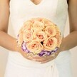 A softly lit, chest-level close-up shows a bride standing calmly, her white lace wedding gown forming a gentle, creamy backdrop to the bouquet she cradles. The bodice of the dress is detailed with fine lace and a modest V-neckline, while the skirt falls in smooth, elegant folds, catching the light like silk. In her hands rests a perfectly rounded bridal bouquet, its heart composed of tightly clustered roses in tender shades of peach, apricot, and blush pink. Each rose is full and velvety, petals curling inward in layered spirals that suggest quiet romance and care. Circling the base of these warm-toned roses is a distinct ring of ruffled blooms in deep purple with white-edged petals, likely lisianthus, creating a dramatic collar of cool colour that makes the soft roses appear to glow. Tiny sprays of white baby's breath are tucked between the blooms, adding a mist-like texture and a traditional bridal touch. The bride's nails are neatly manicured in pale, understated tones, with just a hint of shimmer on one thumb, echoing the subtle sparkle often seen on wedding mornings in Golders Green's local churches and registry offices. There is no visible jewellery to distract from the flowers; the focus is entirely on the craftsmanship of the bouquet and the serene poise of the bride. The scene feels intimate and timeless, like a quiet pause just before walking out towards the guests gathered from around Golders Green, Hampstead Garden Suburb, and along Finchley Road, all waiting to see this bouquet lead the way down the aisle.