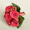 Seen from a high, almost bird's-eye view, this compact bouquet rests against a pale beige, slightly nubby fabric that resembles linen or soft burlap, the gentle texture giving the image a calm and natural backdrop. In the center, several full-bloom red roses-five or six in number-are gathered so closely that their velvety petals overlap, each blossom revealing a perfect spiral heart that suggests classic romance and enduring affection. Nestled between the roses are tight clusters of round, shining hypericum berries in a bright red that edges toward warm orange, their smooth surfaces catching the light and adding an appealing, bead-like texture. Encircling the flowers, wide, dark green tropical leaves have been folded and curled with care, wrapping around the base of the bouquet like a protective shell and giving the design a sleek, slightly contemporary feel. A coordinated red satin ribbon winds around the stems, glimpsed discreetly beneath the foliage, its gentle sheen offering a refined finishing touch that any experienced florist would be proud of. The lighting is soft and even, enhancing the intense reds and deep greens without harsh shadows, making the bouquet appear freshly arranged and ready to be handed to someone special. This arrangement would feel perfectly at home in Golders Green, perhaps carried by a bride walking out of a local registry office, or presented as an anniversary surprise delivered to a cosy flat near Brent Cross. Its overall impression is one of confident elegance and heartfelt emotion, suitable for weddings, romantic milestones, or any occasion where love needs no words.