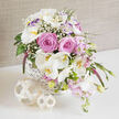 A romantic, garden-inspired floral basket sits beautifully arranged on a glossy white surface, as if ready to be carried into a wedding reception at a boutique venue off Finchley Road in Golders Green. The composition flows from a white wicker basket with an intricate, cross-hatched weave that feels both traditional and stylish. From its brim, a generous cascade of flowers tumbles gracefully, dominated by fully opened blush-pink roses whose layered, velvety petals evoke softness and heartfelt affection. Around and between them, pristine white Phalaenopsis orchids arch outward, their elegant, flat petals and golden centres lending the arrangement a refined, almost sculptural presence reminiscent of high-end hotel décor. White lisianthus blooms, some with gentle purple-tinted edges, mingle with frothy clusters of tiny white gypsophila, creating a cloud-like lightness that keeps the design airy rather than heavy. Smaller blush and soft pink blossoms, suggesting fragrant stock or Sweet William, trail gently, enhancing the sense of movement and giving a feeling of a hand-gathered garden bouquet. A single, slender spike of deep magenta or purple veronica rises playfully from the mix, offering a sleek vertical line and a richer note of colour. Lush green leaves, delicate stems, and a few curling shoots frame the florals, adding depth and freshness, as if they were cut that morning from a nearby Golders Green garden. In front of the basket, two pale, rattan-style decorative balls rest on the tabletop, one slightly larger, their open lattice texture echoing the basket's weave. Bathed in soft, natural light and set against a muted taupe fabric backdrop, this arrangement radiates dreamy sophistication and would feel perfectly at home gracing a bridal table or intimate celebration across NW11.