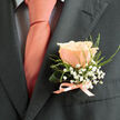 This boutonniere is an exercise in gentle contrast and whispered celebration, crafted to rest comfortably on a groom's lapel or a devoted guest's jacket. A singular peach rose commands the composition with soft, warm petals that spiral inward like the first light of dawn; its coral-pink tones are luminous without being loud. Surrounding the central bloom, delicate sprays of white baby's breath create a cloud-like cushion, their tiny blossoms scattering points of innocence and light. Fine green foliage-slender, slightly serrated leaves with a fresh, early-morning feel-frames the rose and lends structure while keeping the overall silhouette airy and elegant. The stems are bound with a light peach silk ribbon tied into a modest bow, the satin catching a subtle sheen that mirrors an accompanying tie.

Placed against a charcoal suit, the boutonniere's colours glow with added depth; the dark fabric makes the peach and white sing with clarity. Texture plays through the piece: the rose's velvet, the baby's breath's papery fineness, and the foliage's soft resilience invite touch without asking for it. A gentle floral perfume seems present, kind and hopeful-perfect for weddings, anniversaries, or vows exchanged near familiar Golders Green streets like Hoop Lane or Finchley Road. The arrangement is composed with the care of an experienced hand, balancing tradition and personality so that it reads as both ceremonious and affectionate. Lightweight, balanced, and quietly spirited, this boutonniere feels made for moments that matter.