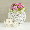 On a pristine white tabletop that subtly mirrors the scene, a carefully crafted wedding centerpiece takes pride of place, radiating a calm yet joyful energy. The flowers form a neat, dome-shaped bouquet, packed with pastel pink roses whose silky petals unfurl gracefully, some edged with a slightly deeper blush that adds depth and romance. Nestled among them, vivid lime-green button chrysanthemums cluster like tiny garden jewels, their compact form and bright colour providing an uplifting contrast to the softness of the roses. Around the base, a scattering of delicate white blooms, similar to small stock or lisianthus, with faint yellow throats, lends a light, airy fringe that gently blurs the boundary between flowers and container. The arrangement sits in a distinctive spherical vase made from white-painted twigs or wicker, meticulously woven into an open, cage-like pattern that feels both handcrafted and contemporary. To the side, two decorative spheres made from fine, pale wood strips echo this woven motif, one slightly larger, creating a pleasing asymmetry and reinforcing the natural theme. The backdrop is an out-of-focus wall in warm beige or taupe, with a subtle texture like woven fabric, providing a cosy, neutral stage for the blooms to glow. The composition evokes the sense of an elegant wedding breakfast table in Golders Green, perhaps not far from the high street or Golders Green station, where a local florist has styled the flowers to bring understated luxury and freshness to the celebration.