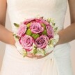 Bathed in the soft glow of Golders Green's late afternoon, a bride's bouquet tells a story of romance, artistry, and nature's harmonious balance. The bouquet's gently rounded form nestles in the crook of her arms, its pastel roses unfurling like petals opening to the morning sun in Golders Hill Park. Those roses, in delicate lavender and dusky mauve, deepen at the rim to brilliant fuchsia-each bloom a miniature sunrise and the heart of wedding-day hope. Encircling this tender core, bright lime-green button chrysanthemums add vibrant life and a textured counterpoint, their compact, dew-fresh petals calling to mind early-spring gardens behind Temple Fortune Lane. Wisps of tiny white flowers-spray roses or freesia-form a halo at the edge, their fragrance hinting at purity and celebration, flecked with bright green buds and feathery leaves that complete the composition. The bride's off-white dress, embellished with lace and gentle folds, forms a luminous backdrop that sets off the bouquet's colours and detail. Her hands, polished in soft pink and touched with glitter, cradle the flowers with anticipation and serenity. The lighting is gentle, caressing every element, from the intricate lace to the lush petals, transforming the bouquet into a symbol of promise and artistry available for bespoke Golders Green weddings-a bouquet that speaks in flowers the language of love and new beginnings.