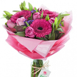 This bouquet radiates an immediate sense of joy and renewal: a hand-tied assembly of vivid fuchsia gerbera daisies with near-black centres, surrounded by softly blushing pink roses at different stages of opening, from tight, elegant buds to full, layered blooms. Small clusters of deep purple-blue statice or similar filler flowers thread through the composition, their papery texture and concentrated colour providing a delicate contrast to the plush petals. Lush green foliage - broad glossy leaves and slender stems - frames each flower, giving the arrangement depth and a fresh, grounded silhouette.

The flowers sit within a clear cylindrical glass vase so the bundled stems are visible below; a length of rustic twine is knotted around the vase neck and a small white rectangular tag hangs from it, printed with a stylised pink-and-green rose logo that identifies the maker. The bouquet is wrapped in layers of sheer, crinkled pink cellophane or tissue that graduates from an almost translucent pastel halo around the blossoms down to a brighter, more saturated pink at the base, catching and scattering soft daylight. The overall scent is gentle and sweet, like an early spring morning in Golders Green when market stalls begin to open - floral, green, and quietly uplifting. As a florist I'd imagine this arranged for birthdays, a thoughtful congratulations, or to lift someone's spirits; its balance of bold gerberas and tender roses makes it equally suitable for lively celebrations and heartfelt, personal gestures across NW11 and nearby Finchley Road.