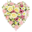 A beautifully detailed heart-shaped floral tribute is shown in full view, resting visually against a spotless white background that allows its tender colours to shine. The heart is generously filled with flowers, leaving no gaps, creating a feeling of embrace and protection. Soft pastel pink carnations form much of the base, their ruffled petals overlapping to give a deeply textured, cushion-like effect that clearly defines the heart's curves and contours. Nestled into this pink bed are several large cream and pale-yellow roses, their spiralling centres and smooth petals bringing a sense of calm, gentle love, and gratitude. Scattered across the arrangement, pure white spider chrysanthemums stand out, each with slender, spiky petals surrounding fresh green centres, adding lightness and intricate detail. Here and there, clusters of deep magenta-pink lisianthus, with softly fluted, bell-shaped blossoms, add emotional depth and a richer hue, like accents of cherished memories. Small sprays of green foliage peek out between blooms, introducing a touch of natural woodland freshness, reminiscent of the leafy streets and gardens around Golders Green. At the pointed tip of the heart, a small pink ribbon can just be seen, subtly suggesting that this design is prepared as a funeral or memorial tribute, ready to be placed at a chapel, crematorium, or graveside. The overall composition is carefully balanced, conveying sympathy, compassion, and enduring love, a fitting floral expression for families in Golders Green seeking to honour a loved one's life with elegance and quiet beauty.