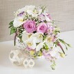 A beautifully composed wedding-style table arrangement rests within a round white wicker basket, its generous mix of pink and white blooms creating an atmosphere of gentle luxury. At the center, large pink roses are fully open, revealing every subtle curve of their spiral petals, their blush tones suggesting romance and heartfelt affection. Surrounding and weaving through them, white Phalaenopsis orchids unfold like porcelain wings, each flower showcasing a bright yellow-orange center that adds warmth and detail to the design. Tucked among these statement blooms, white Lisianthus with softly ruffled petals-and the faintest fringe of lavender-purple on some edges-introduce a tender, layered texture that feels both airy and refined. Pale pink stock flowers arc softly over the basket's rim, giving the arrangement a slight cascade and a sense of movement, as if it were the centerpiece for a reception table in one of Golders Green's intimate venues. Tiny sprays of white baby's breath shimmer between the larger flowers, creating a fine, cloud-like veil that lightens the overall look. Fresh green foliage peeks from within the bouquet and trails gently from the sides, providing contrast and an organic, garden-fresh mood. The woven white basket sits on a pristine white surface, accompanied by two decorative orbs crafted from intertwined pale wooden or rattan strands, echoing the natural weave and adding sculptural interest. Behind, a soft, neutral fabric backdrop-similar to light linen-creates a calm, homely setting that lets the colours and textures stand out. The overall feeling is one of intimate celebration and careful craftsmanship, perfectly suited to weddings, anniversaries, or special gatherings across Golders Green.