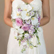 The image shows a waist-level view of a bride in a smooth white strapless wedding dress, her hands gently cradling a cascading bouquet that appears to flow like water. The background is softly blurred in neutral beige tones, allowing the intricate blend of white, pink, and lavender flowers to take centre stage. At the forefront, several large Phalaenopsis orchids unfurl pristine white petals around warm yellow-orange centres, lending an elegant, almost sculptural presence. Clustered between and behind these orchids are rich pink roses and softer, dusty lavender-pink garden roses, their velvety layers suggesting both luxury and intimacy, like a quiet ceremony in a Golders Green townhouse or a reception near Golders Hill Park. Delicate white blooms edged with soft purple, likely lisianthus, peek out from the arrangement, their ruffled petals adding a gentle, romantic rhythm. Wisps of white baby's breath create small, airy clouds, weaving lightness throughout the bouquet and softening every transition between the larger flowers. Slender stems of light lavender stock add subtle height and texture, hinting at a cool, floral fragrance mixing with the crisp scent of fresh greenery. The bouquet narrows as it cascades downward in a refined teardrop shape, echoing the natural line of the bride's dress and drawing the eye towards the hem. Crafted with the care of an experienced North West London florist, this arrangement feels ideal for brides marrying in and around Golders Green, reflecting both modern sophistication and classic romance in every petal.