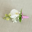 A delicate white orchid boutonniere is laid horizontally on a softly textured, oatmeal-coloured linen background, its calm, neutral tones evoking the quiet elegance of a Golders Green morning. At the centre sits a single Phalaenopsis bloom, its broad, velvety petals a pure, cool white that gently curves outward, cradling a finely detailed throat brushed with soft yellow and a whisper of pale pink. Around the orchid's base, tiny clusters of cloud-like white Gypsophila form a halo of baby's breath, their miniature, rounded blossoms creating an airy, almost misty texture that feels light and romantic. Slender, vibrant green leaves tuck in behind the flowers, their fresh colour offering a crisp contrast against the white petals and the neutral fabric beneath. To one side, a small side stem carries several tightly closed, lime-green orchid buds, their smooth, rounded forms hinting at blossoms still to come and adding a subtle sense of movement and natural growth. The stems are neatly gathered and wrapped in a bright fuchsia satin ribbon, the glossy material tied into a small, precise bow with short trailing ends that catch the light. The pop of vivid pink adds a modern, celebratory note against the tranquil backdrop, ideal for a groom's lapel or prom suit in the Golders Green and Hampstead Garden Suburb area. Gentle, even lighting softly illuminates every petal, bud, and ribbon fold, highlighting the care and craftsmanship behind a floral accessory suited to weddings, civil ceremonies, and elegant celebrations across North West London.