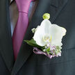 The image captures a meticulously crafted groom's boutonniere pinned to a dark, finely tailored suit, the kind you might see at a smart wedding reception near Golders Green station. At its heart is a luminous white orchid, its petals broad and smooth like satin, unfolding outwards with calm confidence. Around it, slender stems of lilac and clusters of airy white gypsophila nestle among muted sage and silvery-green leaves, creating a gentle halo of colour that softens the strong line of the lapel. A narrow violet ribbon is wrapped neatly around the stems, its satiny finish picking up the lilac tones in the nearby tie, which appears in the background as a soft wash of purple-pink. The palette is serene and contemporary: pure white, cool greens, and dusky mauves working together in quiet harmony. You can almost sense the faint, green fragrance of freshly conditioned flowers, as if they have just been arranged in a nearby Golders Green floral studio and delivered in time for the day's celebrations. Light falls from one side, emphasizing the three-dimensional form of the orchid and casting a slight shadow on the suit, giving the boutonniere a sculptural presence. Every detail-from the cleanly trimmed stems to the careful spacing of each bloom-speaks of intention and affection. It feels perfectly suited to a wedding at a local synagogue or hotel along Finchley Road, an elegant finishing touch that turns a formal suit into a personal, meaningful expression of joy and commitment.