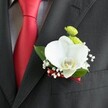 A close-up photograph focuses on a single, meticulously crafted wedding boutonniere resting on the lapel of a deep charcoal suit jacket, the fine wool fabric showing a subtle, woven texture that feels both formal and soft. At the heart of the design sits a large white Phalaenopsis orchid, its broad, velvety petals gently cupped as if in mid-bloom, with a delicately detailed yellow and white lip at the centre that draws the eye like a tiny spotlight. Just above and behind the orchid, two petite, lime-green chrysanthemum button blooms add a refreshing pop of colour, their tightly packed petals creating a cushion-like texture that contrasts with the orchid's smooth surface. Threads of delicate white baby's breath weave through the arrangement, each tiny blossom like a snowflake suspended in mid-air, softening the overall look and giving the boutonniere a cloud-like halo. Rich green leaves frame the flowers, their slightly glossy surfaces catching glints of light and grounding the piece in natural freshness. A slim, deep red satin ribbon binds the stems neatly at the base, perfectly echoing the nearby solid red tie that lies sharply against a light grey and white mini-check shirt, hinting at the careful coordination of a groom dressing for his big day in Golders Green. The light falls from one side, gently highlighting the orchid's curves, the crisp shirt pattern, and the edge of the jacket pocket just beneath, suggesting a poised, composed figure ready to step out for a wedding ceremony. The overall effect is intimate and romantic, a small but meaningful floral accent that feels at once modern, timeless, and lovingly designed for a special celebration in North West London.