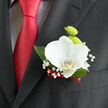 Pinned neatly to a classic black lapel, this boutonnière showcases a flawless white orchid as its serene focal point, the petals unfolding like polished ivory with a gentle, creamy glow at their centre. The orchid's curves feel sculptural and calm, giving the impression of stillness amid the excitement of a wedding morning in Golders Green. Tucked just behind, two petite lime-green button chrysanthemums provide a burst of fresh colour, their rounded forms and tightly layered petals adding playful texture that suggests new beginnings and early spring growth. Encircling the main blooms, sprigs of baby's breath drift lightly outward, each tiny white blossom creating a delicate constellation that softens the edges and adds an almost mist-like effect. A few glossy green leaves slip from beneath, framing the flowers with natural structure reminiscent of the foliage lining the paths near Hampstead Garden Suburb. At the base, a slender ribbon in deep red wraps around the stems, tying them neatly together and mirroring the luxurious red tie worn beneath the suit jacket. This touch of colour adds warmth and romance without overpowering the calm palette of white and green. The boutonnière's proportions sit just right on the lapel-refined yet unassuming, ideal for a groom, best man, or cherished guest gathering for a ceremony close to Golders Green station. Crafted with a florist's eye for detail, the mix of smooth, crisp petals and airy, feathery accents turns this small adornment into a meaningful finishing touch for an elegant North-West London celebration.