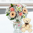 A romantic anniversary gift display is captured on a clean, white, gently reflective surface, where a pastel bouquet in a clear glass jar vase sits side by side with a cream teddy bear. The bouquet is lush but delicate, composed of pale pink roses and peach gerbera daisies with distinctive dark brown centres, framed by crisp white chrysanthemums and touches of lime-green at their hearts. Wisps of white baby's breath weave between the larger blooms, giving the whole arrangement an airy softness that feels like early morning light along the quiet streets of Golders Green. The transparent vase has a homely mason-jar character, its neck wrapped in natural twine tied into a modest bow. Hanging from the twine is a white rectangular tag, decorated with a small pink heart and flowing green lettering that suggests a lovingly handmade or specially curated present. To the right, the teddy bear sits upright, plush cream fur slightly tousled, dark brown bead-like eyes and a small matching nose lending an expression of gentle companionship. A slim dark brown satin ribbon is tied neatly at its neck, mirroring the darker tones in the flowers. Behind them, a softly blurred, light-filled background hints at a window or bright room, keeping the focus on the thoughtful pairing of blooms and bear. It is easy to imagine this arrangement being delivered by a local Golders Green florist as an anniversary surprise, bringing both visual beauty and cosy comfort to a home near the station or tucked off Golders Green Road.