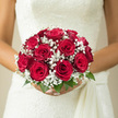 In this image, a bride holds a meticulously arranged bouquet of red roses and white baby's breath, the flowers resting gently against an ivory wedding gown that catches the light like soft satin. Eleven or so crimson roses are clustered tightly at the centre, their petals plush and velvety, opening in spirals that draw the eye inward. Each bloom seems at its peak, neither too tight nor overblown, giving the bouquet a confident, composed presence. Surrounding the roses, delicate sprays of baby's breath form a misty ring of tiny white blossoms, creating a light, airy contrast that recalls the fine lace of a veil or the delicate patterns seen in the shop windows along Golders Green Road. The bouquet is finished with a subtle rim of glossy green leaves, neatly trimmed to keep the shape perfectly round and easy to carry down the aisle of a local Golders Green venue. The colours are bold yet refined: deep scarlet against clean white, softened by hints of fresh green, much like a burst of colour against the quiet tones of nearby Hampstead Garden Suburb. One can almost sense the fragrance-a mix of sweet rose perfume and the gentle, almost powdery scent of gypsophila-surrounding the bride as she steps out for photos in nearby Golders Hill Park. Every element of the bouquet speaks of intention and romance, from the symmetry of its domed silhouette to the way the baby's breath nestles into the spaces between the roses, turning a traditional red-and-white palette into something intimate, emotional, and unforgettable for a Golders Green wedding day.