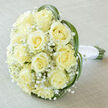 Captured with soft, diffuse light, this bridal bouquet is a study in understated luxury: a rounded cluster of about fifteen to twenty creamy-yellow roses that form a plush, domed centre, each bloom unfurling to reveal perfectly spiralled hearts. Between the roses sit fine sprays of white Gypsophila, their tiny blooms creating a misty, cloud-like texture that lifts the heavier rose heads and imparts a feeling of lightness. Around the outside, several slim, bright-green leaves - likely bear grass or lily grass - arc and curve with purposeful elegance, tracing a contour that both contains and highlights the floral mass. The stems are visible at the base and are meticulously wrapped in clean white satin ribbon, the finish smooth and reassuring to the touch, suited to a bride's careful hold. The bouquet has been placed on a pale taupe linen backdrop that provides a warm neutral contrast, while the soft lighting subtly reveals the faint variations of cream, ivory, and pale yellow in each petal. Working from our studio near Golders Green Road and Temple Fortune, I often recommend this composition for morning or early-afternoon ceremonies where delicate scent and classic lines set the tone; it reads of serenity, fidelity, and quiet joy. Whether chosen for a wedding, a renewal of vows, or as a thoughtful anniversary gift delivered within Golders Green and neighbouring Hampstead, this arrangement balances floral craftsmanship with a reassuringly timeless aesthetic.