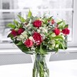 A lush, romantic bouquet of red roses, red carnations, white alstroemeria, and clouds of delicate baby's breath stands proudly in a clear glass vase on a spotless white table, bathed in soft daylight from a large window. The rich crimson roses are the stars, some fully open with velvety petals curling outward in layered spirals, others still in elegant bud form that promise more beauty in the days ahead. Nestled closely around them are ruffled carnations in the same deep red hue, their densely packed petals adding texture and traditional charm, like the kind of flowers you might see in long-loved family homes across Golders Green. Threaded between the red blooms are clusters of white alstroemeria, some tightly closed and green-tipped, others just beginning to open, revealing slender, trumpet-shaped petals with faint markings and a fresh, almost crisp look. Fine sprays of white gypsophila, or baby's breath, drift through the bouquet like a gentle mist, softening the bold colours and catching tiny glints of light. Surrounding this floral heart is a collar of glossy, deep green leaves that anchor the design and give it a sculpted shape, while slimmer, lighter green foliage winds upward, adding movement and a hint of woodland freshness. The stems are clearly visible through the cylindrical vase, neatly cut and submerged in clear water that reflects the brightness of the tabletop. Behind, the view through the window is muted and slightly blurred, like an overcast day in North West London, ensuring every eye is drawn to the vivid reds, crisp whites, and layered greens. The overall feeling is one of thoughtful celebration and tender affection, fitting for anniversaries, romantic surprises, or simply bringing warmth into a Golders Green living room or office.