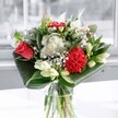 A contemporary glass-vase arrangement of rich red roses, ruffled crimson carnations, creamy white lilies, and daisy-like chrysanthemums is captured in soft daylight, creating a feeling of clear, cool freshness. The focal red rose sits slightly above the rest, its coral-red petals spiralling open with velvety depth, while the carnation beside it has frilled edges that look almost like ripples of silk. Around these passionate tones, clusters of small, snowy white blooms and button-shaped chrysanthemums form a gentle halo, their tiny petals giving the bouquet a cloud-like lightness. Several lily buds, still closed and elegant, rise through the design like pale ivory spears, promising future blossoms and adding a sculptural, modern touch. At the base, deep emerald leaves are folded and wrapped in wide, polished arcs that cradle the flowers and emphasise the arrangement's clean, architectural lines. The clear glass vase reveals neat green stems in the water, underlining the sense of freshness and careful craftsmanship you would expect from a seasoned florist in Golders Green. Natural light, similar to what you might find streaming through a front-room window off Finchley Road, skims across every petal and leaf, highlighting textures from satin-smooth to softly feathery. The overall impression is of an uplifting, balanced bouquet, suitable for a birthday, a quiet celebration at home, or as a simple gesture to bring brightness and calm to someone's day in and around NW11.