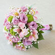 Set against a pale, stone-grey linen backdrop, this lush wedding bouquet seems to float in soft daylight, every bloom carefully gathered to create a rounded, almost domed silhouette. The eye is first drawn to the many lavender roses, their petals layered in tight, velvety swirls that give the design its luxurious, romantic core. Between them, elegant stems of alstroemeria unfurl in shades of blush pink and creamy white, each petal streaked with warm yellow and fine, speckled markings that add an artist's touch. Scattered throughout are neat, spherical button chrysanthemums in bright chartreuse green, their compact, bobbled heads bringing a burst of energy and a modern garden feel. Delicate sprays of white gypsophila weave gently between the larger flowers, creating a veil-like softness and filling the tiny gaps with a cloud of miniature blossoms. The stems are bound firmly together with a wide sash of satin in a pink tone that echoes the roses, the fabric smooth and lustrous beneath the hand. A slender garland of pearls is wrapped over the ribbon, catching the light with each curve, while the trimmed stems extend beyond the binding in a tidy, natural cluster. The bouquet feels freshly made, as though a local florist near Golders Green station has just finished tying it for a bride on her way to a reception near Hampstead Garden Suburb. It radiates the gentle promise of spring, equally at home in wedding photos, engagement celebrations, or as a thoughtful, hand-delivered surprise to mark a cherished milestone within the community.