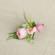 This image shows a refined pink wedding boutonniere carefully arranged as if awaiting collection from a trusted Golders Green florist before a ceremony at a local synagogue or registry office. At its centre is a fully formed pink rose, the bloom opening just enough to reveal its spiralled heart without losing its poised, rounded shape. The petals appear velvety and cool, tinted with an ombré of blush and rose-petal pink, catching soft light that traces their gentle curves. Cradling the central flower are a few pale alstroemeria heads, each petal dusted with fine linear markings and leading into mellow yellow centres that lend a delicate brightness. Between these, airy sprigs of white gypsophila weave in and out, their tiny, round blossoms forming a halo that softens the overall silhouette. Fine green foliage peeks from behind and alongside-slender leaves and feathery fronds that introduce texture and a fresh, garden-like energy, reminiscent of early walks through nearby Golders Hill Park in spring. The stems are trimmed short and bound together with a satin-finish pink ribbon, wrapped in tight, even coils that gleam subtly under the light, suggesting both formality and romance. Everything rests on a subtly patterned, neutral fabric, which offers a gentle contrast to the blush tones and highlights the craftsmanship involved. You can imagine the cool touch of the ribbon, the slightly bouncy resilience of the greenery, and the faint fragrance of the rose mingling with crisp air before a ceremony. Designed for weddings, proms, or important family gatherings around Golders Green, this boutonniere feels like a small, wearable keepsake of tenderness and celebration.