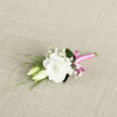 On a softly textured, neutral fabric background, a finely crafted boutonniere rests as if waiting to be pinned for a wedding ceremony in Golders Green. The focal point is a luminous white lisianthus, fully open and plush, with layers of gently ruffled petals unfolding in a rounded, cloud-like shape. There is a quiet luxury in the way the creamy tones transition from a slightly deeper centre to the almost translucent outer edges, giving the bloom real depth. Tucked close at its side are two lisianthus buds, one upright and one leaning inward, their smooth, tapered forms suggesting new blooms on the verge of unfurling. Between them, airy sprigs of baby's breath weave in and out, their tiny white flowers like scattered stars, adding a lace-fine texture without overwhelming the central blossom. Slender green stems and grass-like foliage extend from the base, introducing a sense of movement and natural flow, as though caught in a light breeze on nearby Finchley Road. The stems are expertly aligned and tightly wrapped in a glossy, bright pink satin ribbon that runs the full length of the grip, finished with a small, perfectly formed bow. The ribbon's vibrant magenta-pink hue adds a pop of joyful colour and a subtle contemporary twist to this otherwise classic white-and-green palette. Every element is balanced so the boutonniere feels light, elegant, and heartfelt, ideal for the groom or groomsmen at a local synagogue wedding or civil ceremony in Golders Green, reflecting a blend of tradition, tenderness, and modern style.
