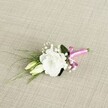 A delicate wedding boutonnière rests at a gentle angle on a soft, oatmeal-coloured fabric that looks like finely woven linen, its subtle texture catching the light and giving the scene a calm, Golders Green morning feel. At the heart of the design is a single white lisianthus bloom, its ruffled petals fully open and layered like fine silk, forming a rounded cup of soft ivory and pure white. Just below, two slender lisianthus buds in creamy pale green lean in close, suggesting flowers that will open later in the day, much like a celebration unfolding from a quiet ceremony at Golders Hill Park to an evening reception nearby. Tiny clusters of white gypsophila are scattered through the arrangement, their cloud-like blossoms adding an airy, romantic fullness around the focal flower. Slender blades of deep green, grass-like foliage extend gracefully towards the lower left, giving movement and a slightly wild, garden-picked touch, while a broader, darker green leaf tucked behind the bloom anchors the composition and adds depth. The stems are neatly bound at the base with a satin ribbon in bright fuchsia pink, tied into a small, jaunty bow that provides a playful burst of colour against the muted background and classic white-and-green palette. The soft, even lighting highlights each petal edge, each tiny gypsophila floret, and the gentle sheen of the ribbon, creating a sense of freshness as if this buttonhole has just been prepared by a local florist for a groom or groomsman getting ready off Finchley Road. The overall impression is of understated elegance with a modern twist, perfect for weddings, proms, or black-tie events around Golders Green, where a single refined detail can quietly express personality and joy.