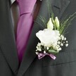 A sharply focused close-up of a groom's lapel shows a dark charcoal grey suit jacket framing a single, exquisitely crafted white lisianthus buttonhole, the kind of refined detail often seen at elegant Golders Green weddings. The wool suit has a subtle textured weave that catches the light softly, with a crisply pressed lapel leading the eye straight to the floral work. At the heart of the boutonnière, the lisianthus bloom is fully open, its layered petals ruffled and velvety, a creamy white that feels almost luminous against the deep grey fabric. Just above, two slender ivory buds rest in quiet anticipation, their tips still gently closed, suggesting flowers that will unfurl later in the day. Wispy sprays of baby's breath surround the main bloom like a light, cloud-soft halo, each tiny white floret adding airy volume without overwhelming the central flower. Fine, linear blades of greenery arch outward, giving movement and an almost ethereal lift to the composition while keeping the overall look clean and modern. At the base, a narrow mauve ribbon is wrapped and tied with care, its soft pinkish-lavender tone echoing the nearby silk tie, which appears in a vibrant magenta shade between the jacket lapels. The tie's smooth sheen contrasts with the patterned white and grey shirt beneath, bringing cohesion and quiet drama to the ensemble. The whole scene feels like a still moment before a ceremony, perhaps in a sunlit registry office off Finchley Road, the fresh, green-and-white scent of the flowers offering a calm counterpoint to the excitement of the day. The image captures polished tailoring, meticulous floristry, and a sense of hopefulness woven into formal attire.