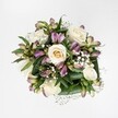 A compact, hand-tied bouquet is viewed directly from above, the flowers laid out like a soft rosette against a pure, bright white background that makes every detail easy to see in the mind's eye. At the heart of the arrangement sit creamy white roses, some still cupped and shy, others fully opened so that their layered petals spiral outwards, revealing buttery ivory centres. Threaded between these roses are stems of alstroemeria, or Peruvian lilies, their narrow buds and gently opening blooms washed in mauve, lavender, and soft berry-pink, with delicate green streaks and freckles that give each petal a painterly look. Fine sprays of gypsophila weave through the bouquet, tiny white clouds of baby's breath that soften the outlines of the larger flowers and add a romantic, floating lightness. All of this is cradled by a collar of deep, glossy green leaves, neatly folded and curved to frame the blooms like polished satin. The lighting is clean and even, as if captured in a calm studio moment somewhere just off Golders Green Road, allowing the textures and subtle colour shifts to stand out with quiet clarity. The bouquet feels fresh, cool, and composed, ideal for imagining as a wedding posy, a thank-you gift, or a gentle get-well gesture delivered to a home near Golders Hill Park. The overall mood is elegant yet approachable, reflecting the careful hands of a local florist who understands how to balance structure, softness, and lasting freshness in every stem.