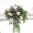 This image shows a softly lit sympathy bouquet that radiates quiet composure and care, much like a comforting presence in a Golders Green home. The flowers are gathered in a tall, clear glass cylinder vase, where the straight green stems can be seen clearly through the water, adding a sense of freshness and authenticity. Above the rim, creamy white roses form the main focus, their petals layered and velvety, opening in serene, rounded blooms that seem to rest gently against one another. Between the roses, alstroemeria blooms and buds rise gracefully, their slender petals brushed with tones of purple, lilac, and pale green, lending a cool, tranquil touch of colour that never distracts from the bouquet's peaceful feeling. Fine sprays of white baby's breath are threaded throughout, appearing like tiny stars scattered among clouds, softening the overall shape and giving the arrangement a light, gossamer quality. The foliage is a rich, natural green, with glossy leaves and delicate stems that cradle the blossoms, adding structure and a quiet sense of life and renewal. Everything sits against a very bright, softly blurred white background, similar to a bouquet placed near a sunlit window overlooking the tree-lined streets around Golders Green station. This brightness makes the whites look particularly clean and pure, while allowing the purples and greens to glow gently. The bouquet feels composed and respectful, perfect for sending sympathy, marking a memorial, or offering a thoughtful gesture of support when someone nearby is grieving and in need of a simple, beautiful expression of care.