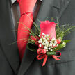 This image shows a refined boutonniere designed for a wedding or formal celebration, resting against the textured lapel of a deep charcoal suit, like you might see at an intimate ceremony near Golders Green station. At its heart is a single pink rose, full and luminous, with petals that curve outward in soft, velvety layers. The shade of pink is rich but not overpowering, suggesting romance and gentle confidence, similar to the blush of sky you catch over nearby Golders Hill Park at sunset. Surrounding the rose, clusters of tiny white gypsophila blossoms bring a light, airy touch, their star-shaped flowers forming a delicate cloud around the central bloom. Nestled among them are small, shining red hypericum berries, perfectly round and jewel-like, their glossy skins adding depth and a playful burst of colour. Slender strands of fresh green foliage peek out from behind, giving the arrangement a natural, slightly wild edge, as though freshly gathered from a well-loved North London garden. A single, smooth green leaf has been shaped into a graceful loop at the base, its polished surface catching the light, providing structure and a modern, sculpted line. The stems are bound together with a narrow red ribbon, tied with precise, tidy turns that speak of an experienced florist's careful touch. The composition is balanced and thoughtful, carrying an air of hopeful anticipation-ideal for a groom, usher, or prom guest wanting a classic yet contemporary floral accent crafted by local hands familiar with the tastes and traditions of Golders Green.