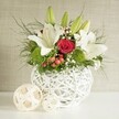 A refined floral arrangement is presented as though ready to grace a wedding table in Golders Green, poised on a clean white surface against a soft, light beige wall. The flowers are gathered in a round white vase with an intricate open-weave pattern, its interlaced strands creating a basket-like texture that feels both modern and organic. Rising from this sculptural base is a lush bouquet in a timeless white, red, and green palette. Several white lilies take centre stage: some are fully open with wide, star-shaped petals and golden stamens, others are still enclosed in elegant green buds that hint at the fragrance yet to come. Among them glows a central red rose, its velvety petals tightly coiled into a perfect spiral, surrounded by other deep red roses that intensify the romantic feel. Bright green, pom-pom-style blooms-likely green dianthus or chrysanthemums-add a lively burst of colour and a pleasing, tufted texture, standing out among the smooth lily petals. Airy clusters of white baby's breath drift throughout the arrangement, softening the composition like a fine mist. Small groups of red berries tuck between the flowers, offering a subtle festive touch that would suit winter celebrations or intimate dinners along Golders Green Road. The bouquet is framed with assorted greenery, from wispy, fern-like tendrils to broader, darker leaves that cascade naturally over the rim of the vase, giving it a sense of movement and fullness. To the left, two decorative woven spheres in light, natural tones sit on the table, echoing the vase's design and completing the coordinated, stylish scene, ideal for wedding receptions and special occasions across the NW11 area.