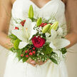 The image shows an elegant bridal bouquet cradled against the lace bodice of a wedding dress, the fine pattern of the fabric framing the flowers like delicate embroidery. At its heart, a classic red rose sits in full bloom, the petals densely layered and luxuriously dark, drawing the eye with its depth of colour and promise of romance. Surrounding it, several large white lilies open wide, their creamy petals slightly curved at the tips, catching the light with a gentle sheen that contrasts beautifully against the matte texture of the lace. The stamens, tinged with warm amber, hint at the soft, sweet fragrance that often fills synagogues and ceremony rooms around Golders Green. Tiny white gypsophila blossoms weave through the design, their cloud-like clusters lending a dreamy softness and bridging the transition between bold red and pristine white. Bright red berries add playful pops of colour, round and glossy, echoing the warmth of family celebrations in nearby Temple Fortune cafés after the vows. Sprigs of rich greenery, some velvety and moss-like, others smooth and leaf-shaped, form a lush base that recalls the leafy avenues off Golders Green Road. Fine, arching grasses give the bouquet an airy, almost ethereal outline, as if the flowers are lightly floating rather than held. The composition feels thoughtfully balanced, neither too formal nor too wild, capturing the emotional mix of excitement, serenity, and hope that defines a wedding day. It's the kind of bouquet a local Golders Green bride might carry from a quiet home on Hoop Lane to a joyful reception filled with laughter, music, and shared memories.