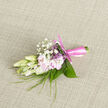 Set against a neutral, softly woven fabric, this small boutonnière appears like a carefully composed poem in cream, pink, and green. At the centre, creamy freesia-style buds are caught in the moment just before they fully open, their petals crisp yet delicately translucent, hinting at the fragrance within. Nestled beside them are pale pink blooms that unfold with gentle curves, each petal velvety and light, creating a subtle play of shadow and softness. Tiny stars of baby's breath hover above, forming delicate clusters that resemble miniature clouds drifting over the rooftops of Golders Green on a calm day. Fine, blade-like greenery arcs through the design, offering slender lines that add structure and a sense of grace, while slightly broader leaves give a reassuring, leafy backdrop, deep green and glossy. The whole arrangement is gathered tightly, the stems neatly aligned and wrapped with a satiny pink ribbon that circles them with precision. The knot is small, firm, and perfectly tied, its soft sheen catching the natural light as if it were a ribbon chosen moments before a ceremony at a local synagogue or registry office near Golders Green station. The imagined scent is subtle yet evocative-sweet, green, and fresh, the sort of fragrance that lingers lightly on a jacket after an evening reception on Finchley Road. Crafted as a wedding or special-occasion lapel piece, this boutonnière feels personal and intimate, adding a gentle touch of colour and nature's artistry to the wearer. It expresses tenderness, attention to detail, and the quiet joy that often accompanies cherished events in the neighbourhoods around North West London.