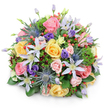 A lush, rounded bouquet sits against a crisp white backdrop, its domed silhouette and soft shadow giving the composition a sculpted, three-dimensional presence. At the heart are garden roses in soft blush pink, warm butter yellow, and gentle peach - their layered petals curling in tight spirals and fuller, open heads that suggest different moments of unfolding. Scattered like punctuation across the surface are star-shaped clematis in pale lavender and near-white, each with a bright yellow eye that lends an airy, garden-wild note to the design. Cool, architectural contrast comes from a handful of blue Eryngium (sea holly); their metallic-blue, spiky heads and bristled collars provide an almost sculptural edge against the roses' softness. Deeper tones are woven through by ruffled purple lisianthus, some gently unfurling, others held in poised buds, while clusters of glossy red Hypericum berries add cheerful, jewel-like accents. The bouquet is nestled in a rich tapestry of greenery: broad glossy leaves form a verdant base, feathery sprigs add depth and movement, and darker burgundy filler foliage peeks through to create layered texture. Light grazes the petals, catching dew-like highlights and lending a fresh, natural glow that suggests a cool morning - the scent imagined as soft, sweet, and garden-fresh. This arrangement reads as both romantic and modern, crafted for occasions that range from celebration to quiet appreciation; it would make a refined table centerpiece or a heartfelt gift for someone who loves English-countryside charm in an urban setting. The bouquet's balance of classic roses, delicate clematis, striking sea holly, and vivid berries demonstrates confident floristry and a thoughtful sense of colour, texture, and scale evocative of neighbourhood markets and green spaces around Golders Green.
