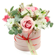 A charming hatbox bouquet in soft pinks, whites, and fresh greens is presented against a clean white background, giving the flowers a spotlight as clear as a bright morning over Golders Green. The container is a smooth, cylindrical box in gentle pastel pink, its matte finish suggesting a luxurious yet understated gift. Around the box runs a slender coral-red satin ribbon, tied into a neat bow at the front, with a cream gift tag hanging delicately from the knot, lettered in green script as if ready to carry a personal message from someone nearby on Golders Green Road.
The floral arrangement rises above the rim of the box in a graceful dome, lush and rounded, with no harsh angles. A prominent bi-colour rose at the front draws attention: its outer petals are pale blush, gradually deepening to a rich pink swirl at the centre, evoking warmth and romance. Surrounding this focal bloom are clusters of white daisy-like chrysanthemums with bright green centres, their crisp petals bringing clarity and brightness reminiscent of suburban gardens in NW11. Interlaced with them are white alstroemeria, their slender, trumpet-shaped petals carrying delicate speckles inside that add quiet refinement.
Dotted through the arrangement are smaller flowers in vivid fuchsia and deep pink-perhaps spray roses or lisianthus-that provide lively pops of colour and a playful accent against the softer tones. A generous selection of greenery, including rounded silvery eucalyptus leaves and darker foliage, frames the blooms, while tiny green buds lend a sense of freshness and new beginnings. The overall impression is of a thoughtfully curated, modern hatbox gift, perfect for same-day delivery to mark birthdays, anniversaries, or simple gestures of appreciation across Golders Green and neighbouring streets off Finchley Road.
