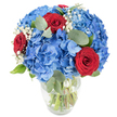 A luminous, hand-arranged bouquet sits in a clear glass vase on a clean white surface, the kind of composition a local florist would pause to perfect. The arrangement is anchored by voluminous clusters of sky-blue hydrangeas whose many small petals form a cloud-like mass, cool and soft in appearance as if they were still holding morning rain. Woven through that blue cloud are several deep, velvety red roses - their petals tightly furled into luxurious spirals that read as declarations of passion against the cool backdrop. Tiny sprays of white baby's breath drift between the larger blooms like flecks of light, lending a delicate, ethereal lace that softens the overall silhouette. Rounded, muted-green eucalyptus leaves and fresher green foliage are threaded through the bouquet, their matte surfaces and subtle herbal scent balancing the sweetness of the flowers and adding sculptural depth. The vase has a gently rounded base and a tapered neck, its transparency revealing stems immersed in clear water and hinting at the bouquet's freshness and careful preparation. Bright, neutral lighting highlights the saturated blues and reds, while the uncluttered white background lets the colours sing without distraction. As a florist with years working in and around Golders Green, I imagine this arrangement brightening a dining table near Golders Hill Park or sitting on a mantelpiece in a Hampstead Garden Suburb flat, offering a confident, celebratory mood - equally suited to anniversaries, birthday toasts, or a patriotic display - and radiating quiet, refined joy together with the soft herbal scent of eucalyptus.