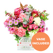 A generously proportioned bouquet brimming with pink roses, ruffled carnations, and cheerful daisy-like blooms sits proudly in a clear glass vase, photographed against a clean white background that makes every colour stand out. The roses, in shades ranging from rich fuchsia to soft, romantic blush, are fully open or just beginning to unfurl, their layered petals hinting at luxury and care. Around them, lavish carnations in coral, peach-pink, and creamy white add rounded volume and a soft, textured surface that looks almost like folded silk. Scattered throughout the arrangement are clusters of lavender and purple chrysanthemums, each small flower marked by a vivid green centre that adds a refreshing burst of brightness. The bouquet is framed and deepened by an abundance of foliage: cool-toned eucalyptus leaves in silvery green, broader dark green leaves, and the occasional reddish-brown accent that gives a subtle hint of autumn warmth, much like the colours you might glimpse in gardens off Golders Green Road as the season shifts. The vase itself is square with gently curved edges, completely transparent so the stems and water are visible, and it is wrapped near the base in a white band decorated with repeating red hearts and the romantic phrase all my love in flowing script, reinforcing that this is a heartfelt gift. In the bottom right corner, an orange circular graphic declares VASE INCLUDED in bold white letters, making clear that this arrangement is ready to place directly on a table or windowsill. The overall feeling is one of affectionate celebration, perfect for sending love, congratulations, or gratitude with a fresh delivery to homes and offices across Golders Green.