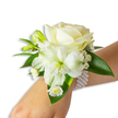 A delicate wrist corsage sits against a clean white backdrop, framed on the fair-skinned wrist of the wearer as the arm extends from the lower right into the centre of the frame. At the heart of the arrangement a velvety, creamy ivory rose opens in soft spirals, its petals catching a cool, diffuse light and lending the piece a gentle, romantic glow. Just below and a little to the left a large trumpet-shaped white bloom - with the slender, slightly ribbed petal texture and pale lime-green throat of alstroemeria - provides a graceful, open silhouette that balances the rose. Small clustered buds of green promise new blooms, while airy sprays of tiny white gypsophila float like whispering clouds around the main flowers. Near the base, petite daisy-like white florets with sunny yellow centres add a sweet, vintage note and a point of bright contrast against the white petals.
Lush, broad deep-green leaves with a waxy sheen frame the composition, their glossy surfaces setting off the pristine whites and suggesting a freshly clipped, garden-picked quality. The blooms are secured by a chic multi-strand pearl bracelet; its round, lustrous beads wrap gently around the wrist, offering a tactile, cool-smooth band that anchors the floral design. The overall effect is one of timeless elegance - a corsage that reads as both classic and intimate, suited to weddings, proms, and formal dances. Imagine the faint, fresh scent of roses and summer stems, and the quiet pride of slipping this onto your wrist before an evening in Golders Green or a photograph beneath the soft light of a nearby park.